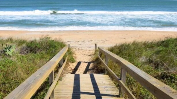 Un camino de madera que lleva a una playa de arena, con olas del océano al fondo.