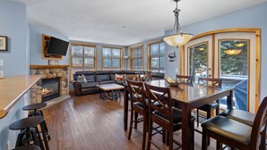 A cozy living room with a fireplace, a dining area with wooden chairs, and large windows with blinds. - Perfectly Located beautiful 4 bedroom townhouse (Sun Peaks)