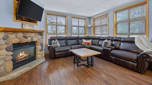 A living room with a stone fireplace, leather sofa, wooden coffee table, and large windows with blinds. - Perfectly Located beautiful 4 bedroom townhouse (Sun Peaks)