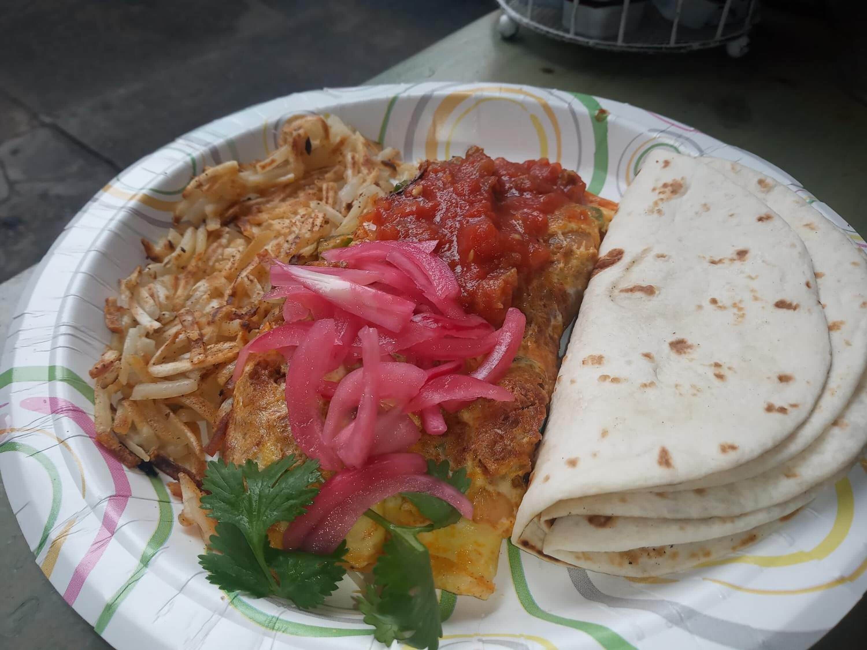A paper plate with a meal consisting of a tortilla, a fried item, a red sauce, and pickled onions.