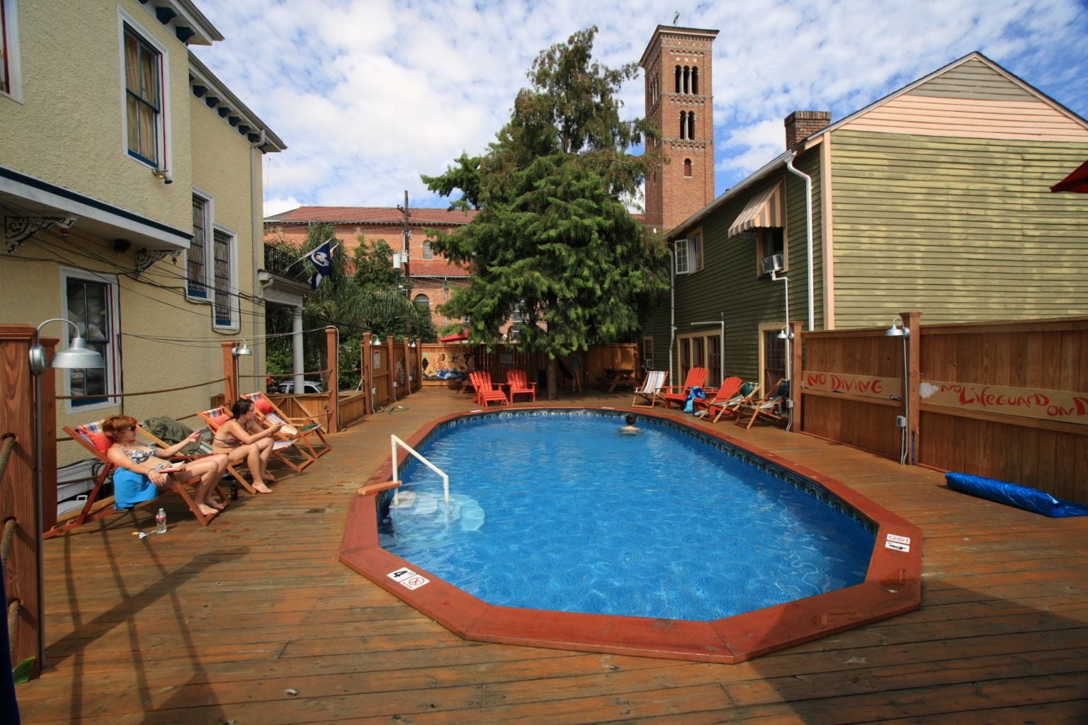 Une piscine rectangulaire entourée d’une terrasse en bois, un bâtiment en briques avec une tour en arrière-plan, et une clôture en bois recouverte de graffitis.