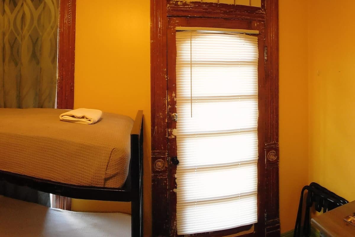 Quarto pequeno com uma cama de solteiro, beliche, espelho, janela com persianas e porta.