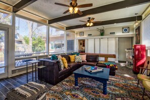 Ein geräumiges Wohnzimmer mit einer dunkelbraunen Ledersofa, einem blauen Couchtisch und einem gemusterten Teppich. Zwei Deckenventilatoren, ein rotes Schränkchen und ein Fenster mit Jalousien sind ebenfalls vorhanden.