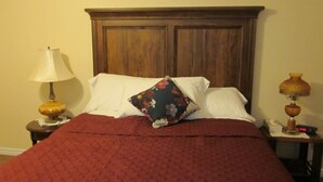 Uma cama com cabeceira de madeira, almofadas brancas, uma almofada florida, um cobertor vermelho e uma luminária em uma mesa de cabeceira.