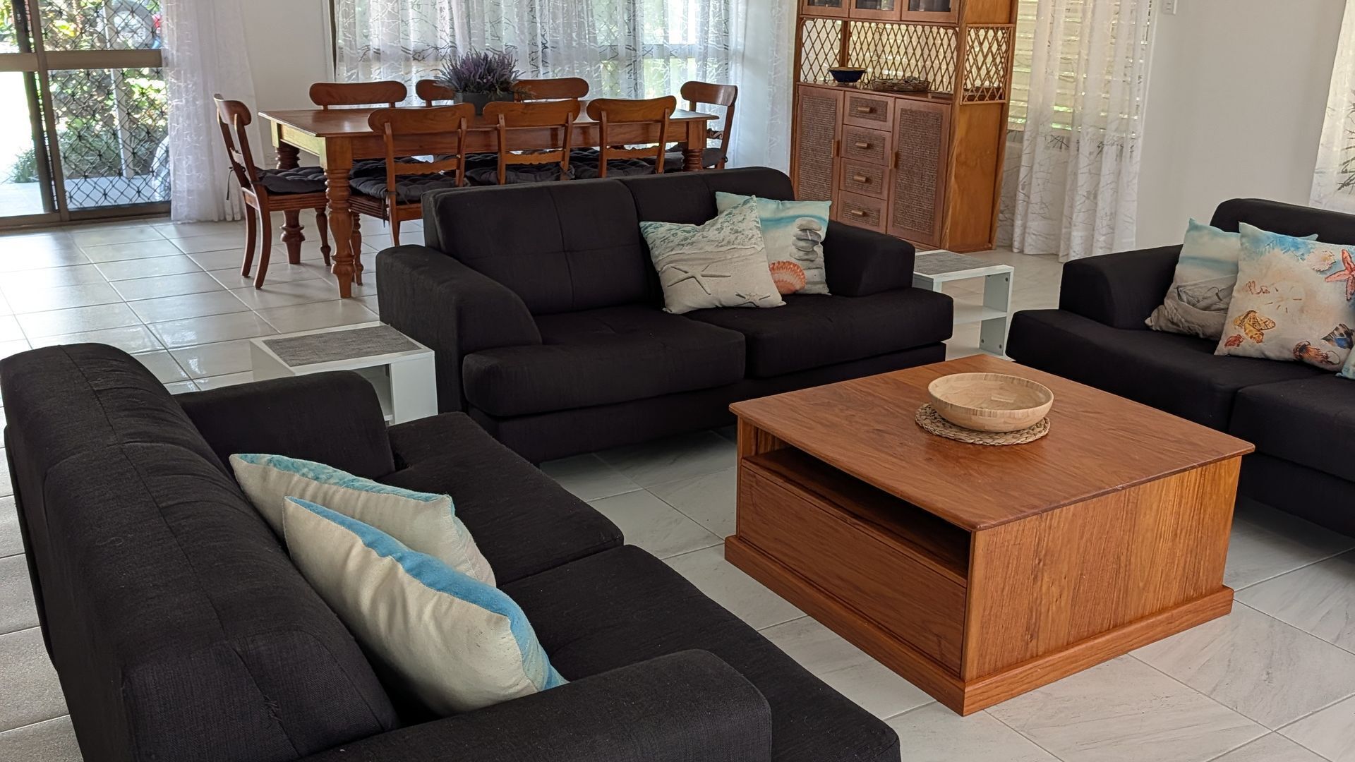 A modern living room with dark sofas, a wooden coffee table, and a dining area with wooden chairs and a table.