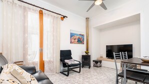 A living room with a sofa, a black armchair, a dining table with chairs, a TV, and a painting of a coastal scene.