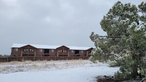 Una cabaña de madera grande de dos pisos con balcón, rodeada de nieve en el suelo y entre árboles.
