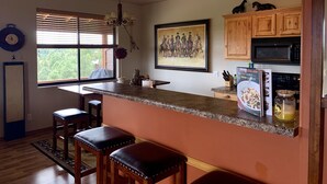 Una cocina con encimera de granito, taburetes de madera, un reloj en la pared y una ventana con persianas.