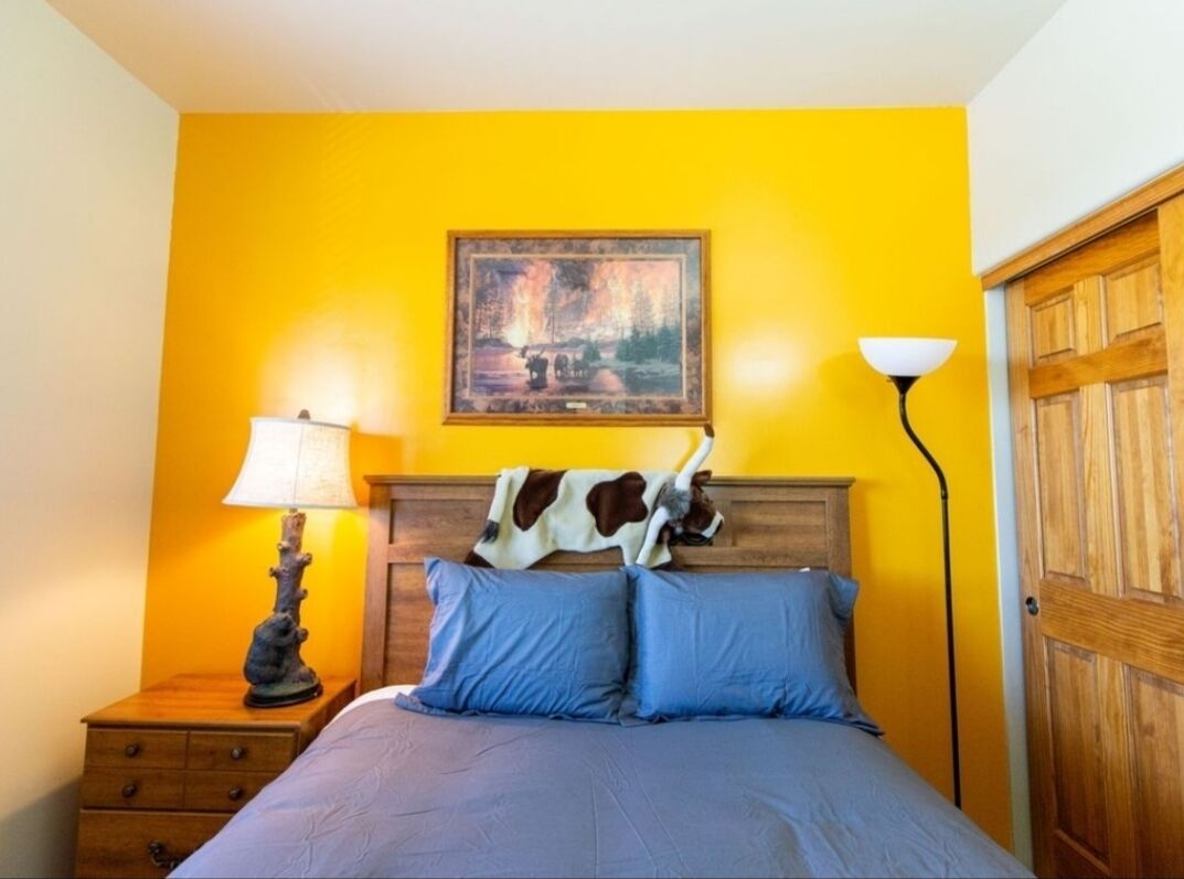A bedroom with a wooden headboard, a painting above the bed, a nightstand with a lamp, and a wooden door.