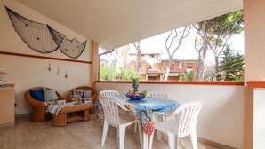 A dining area with a table and chairs, a wicker chair, a small table with a bottle and glasses, and a view of buildings and trees through a window. - Apartment Ancora on the sea (Marina di Grosseto)