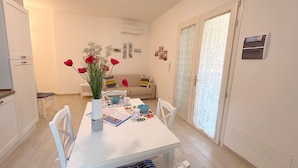 A dining area with a white table, chairs, and a vase with flowers. There is a sofa and a window with blinds in the background. - Apartment Ancora on the sea (Marina di Grosseto)