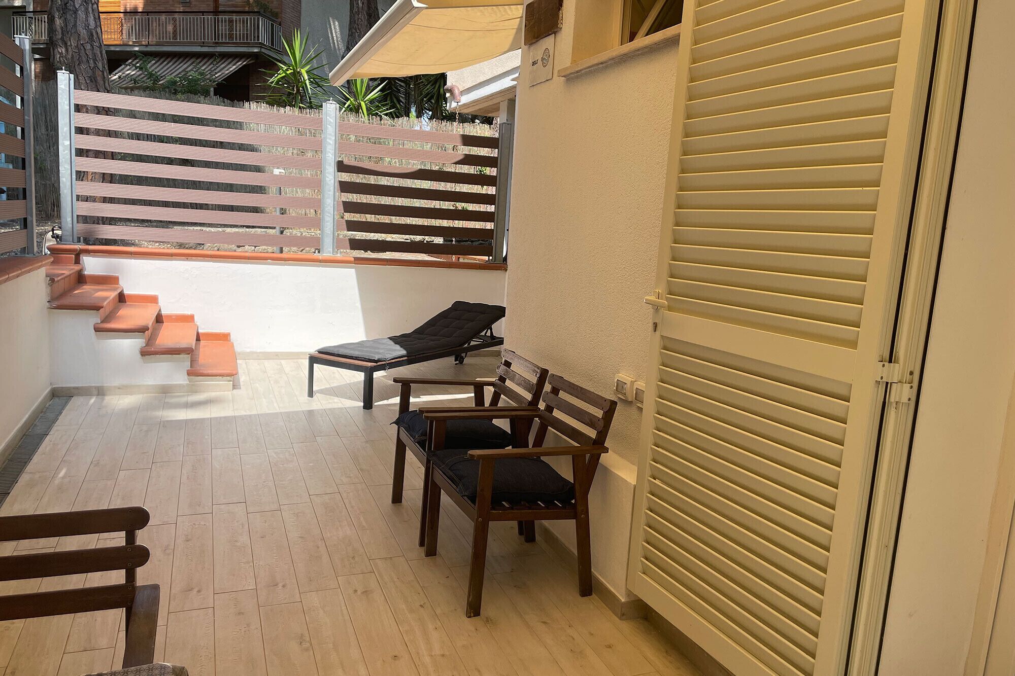 A balcony with a wooden bench, a lounge chair, and a table. There are steps leading to a terrace with a view of a building and plants.