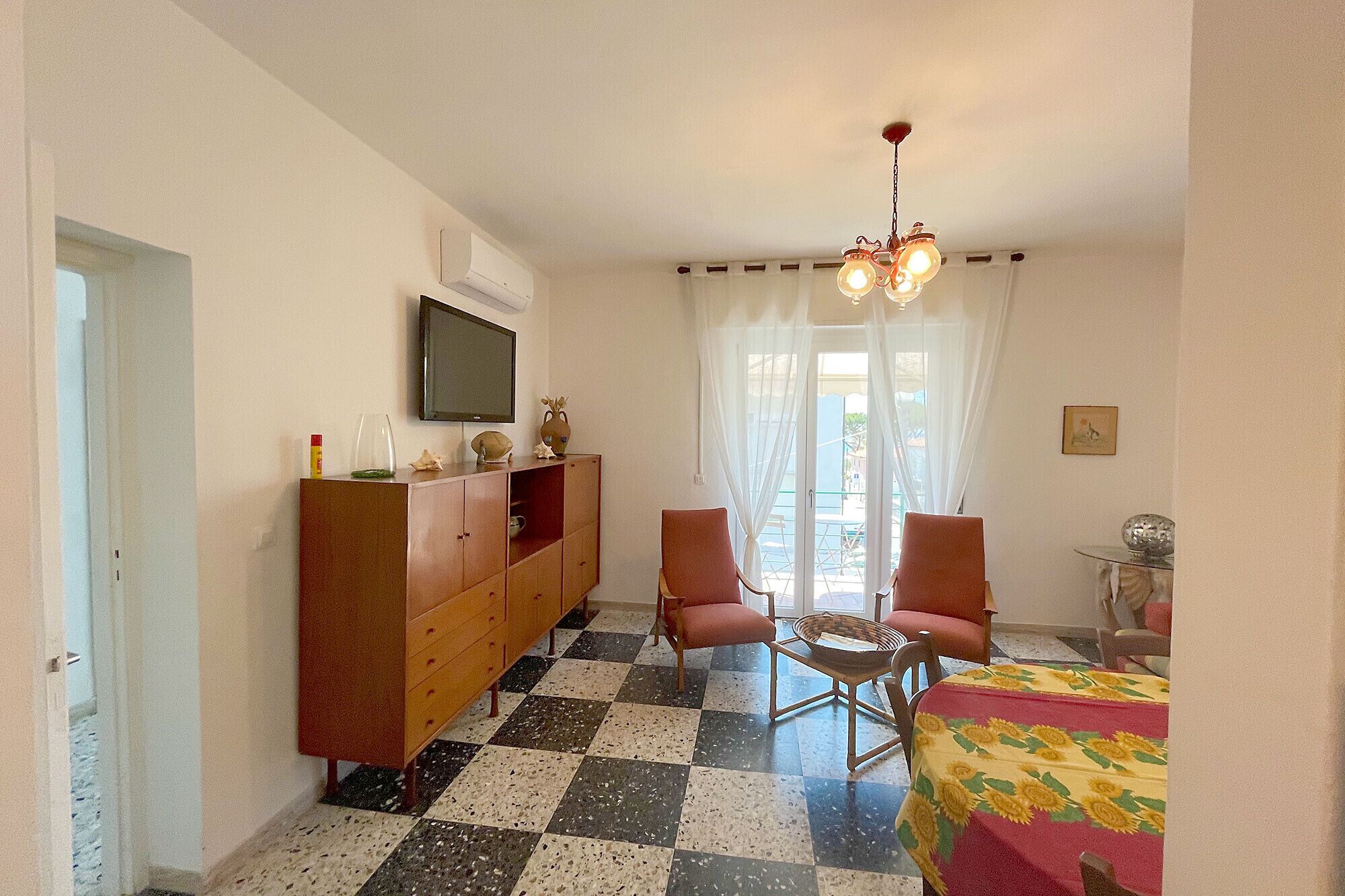 Ein Zimmer mit Schachbrettmuster-Boden, einem an der Wand befestigten Fernseher, einem Holzschrank, zwei Stühlen, einem Tisch mit Sonnenblumen-Tischdecke und einer Chandelüster.
