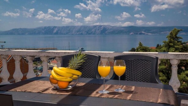 Ein Tisch mit einer Obstschale mit Bananen und Orangen, zwei Orangensaftgläsern und einer Ananas auf einem Balkon mit Blick auf ein Gewässer und Berge.
