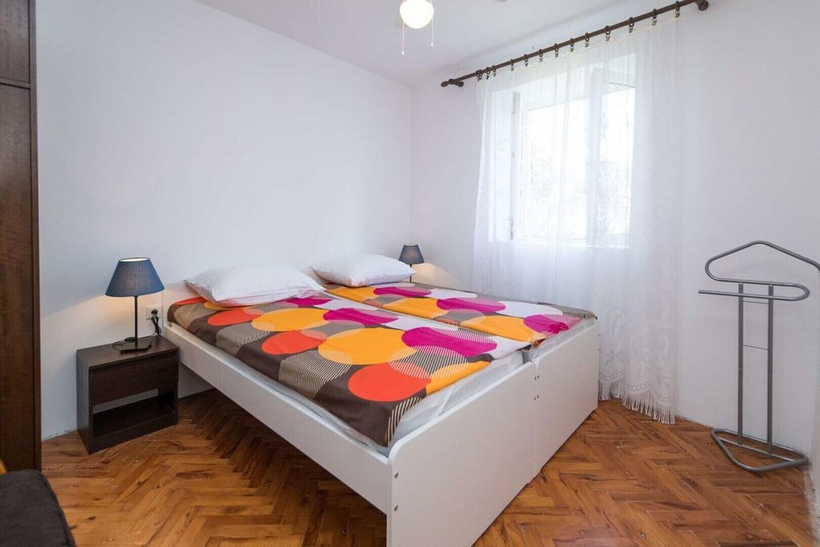 Une chambre avec un parquet en bois disposé en motif chevron, un lit recouvert d’une couvre-lit aux motifs géométriques colorés, une lampe de chevet et une fenêtre ornée de rideaux légers.