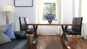 Uma sala de jantar com mesa de madeira, duas cadeiras pretas, um sofá de couro preto com almofada azul, um abajur e uma janela com cortinas.