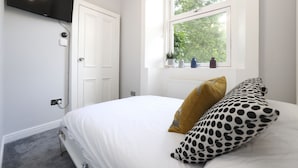 Quarto com cama, janela com vista para árvores, porta branca e televisão de tela plana fixada na parede.