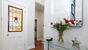 Um corredor com porta branca, um vaso de vidro com flores, uma prateleira de vidro e um espelho na parede.