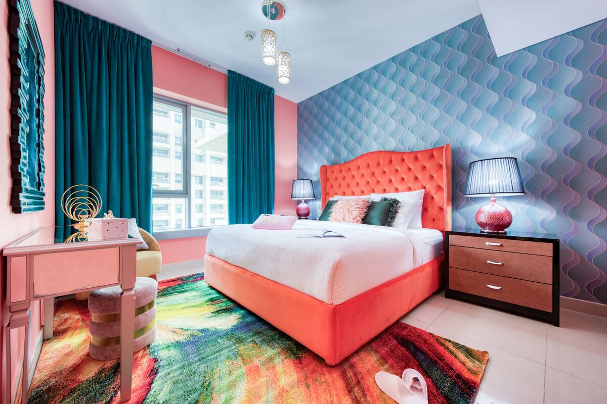 A bedroom with a large bed, a desk, and a dresser. There is a window with curtains, a patterned wall, and a colorful rug.