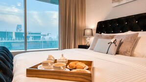 A hotel room with a large bed, a tray of breakfast items, a city view, and a modern headboard.