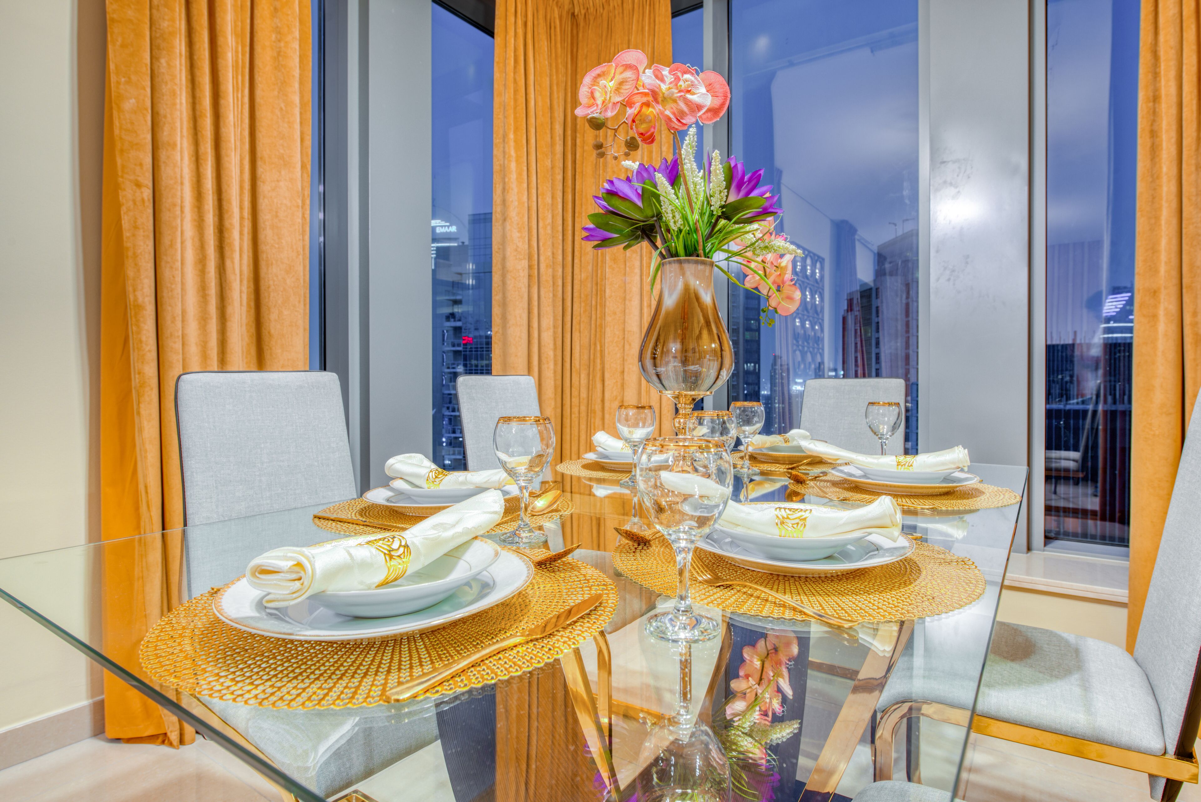 Une table à manger dressée pour un repas, avec un vase de fleurs, des assiettes à bord doré et de la verrerie transparente.