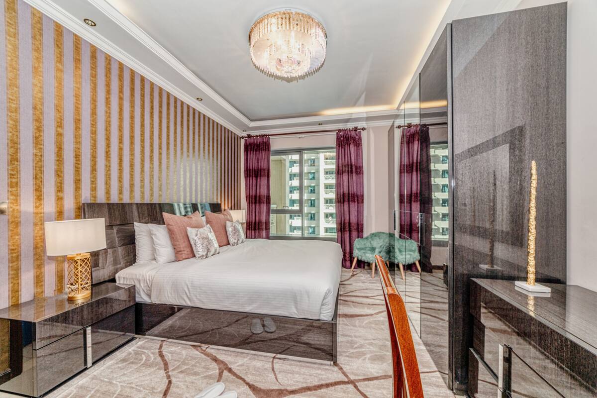 A modern hotel room with a large bed, a chandelier, a TV, and a view of the city.