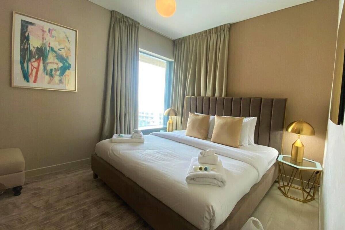 A hotel room with a large bed, a bedside table with a lamp, a window with curtains, and a painting on the wall.