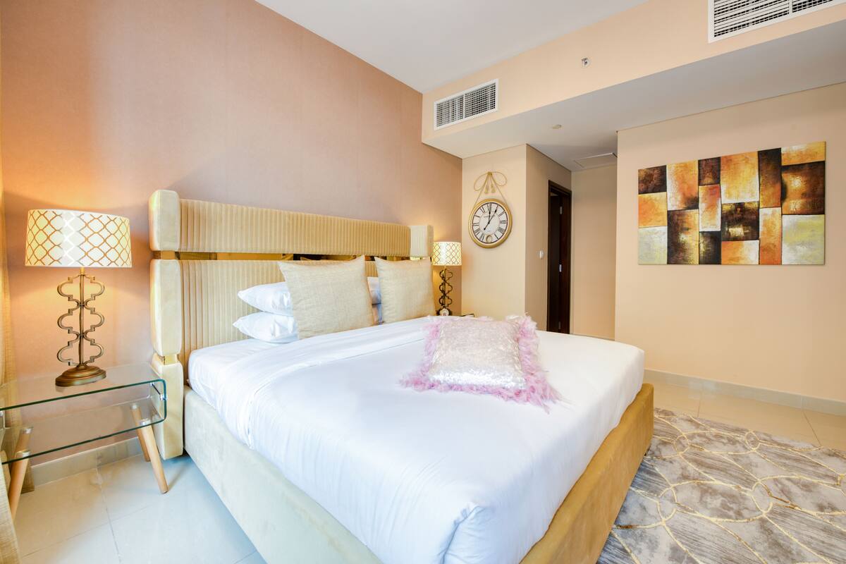 A neatly arranged bedroom with a large bed, bedside lamps, a wall clock, and a modern abstract painting.