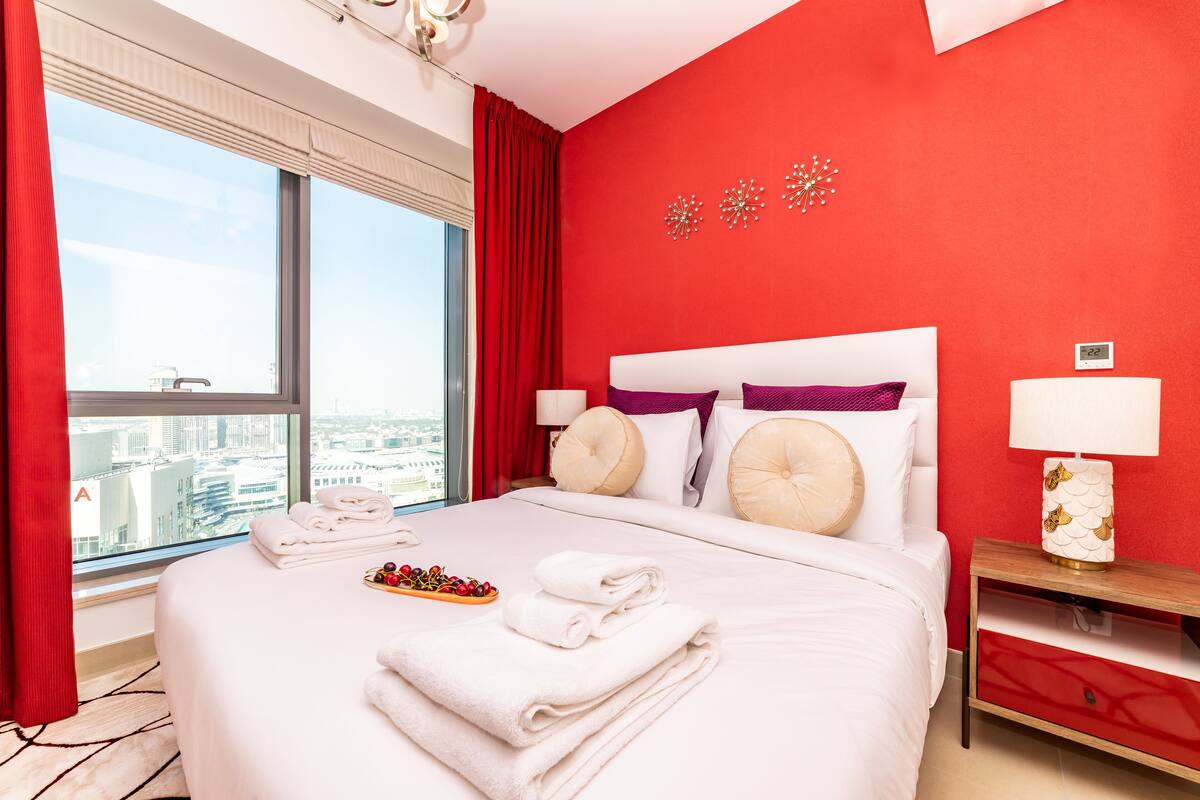 A hotel room with a large bed, red walls, a city view, and a bedside table with a lamp.