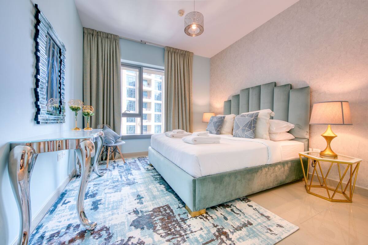 A modern hotel room with a large bed, a desk with a chair, a window with curtains, and a decorative mirror.