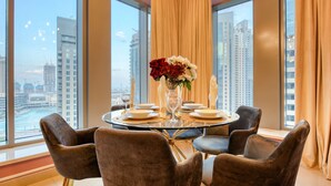 A dining area with a round table set for four, a vase with flowers, and a view of tall buildings and a body of water through large windows.