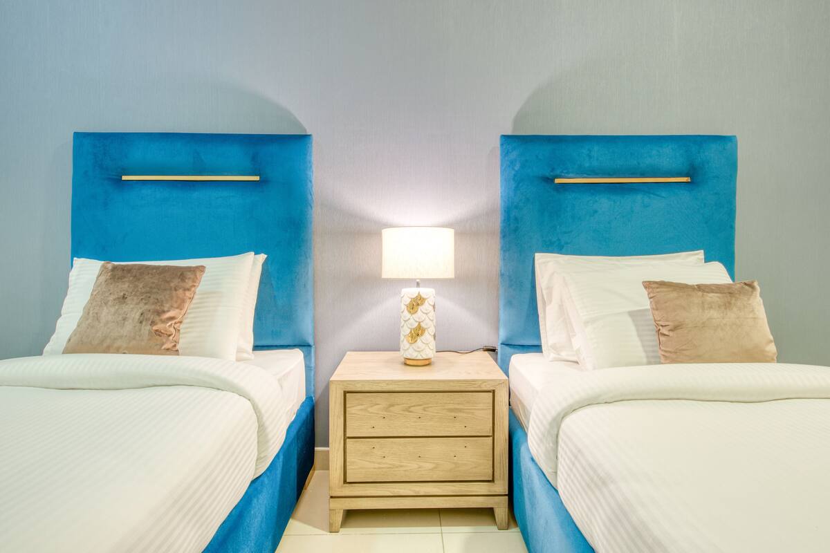 A room with two beds, each with a blue headboard and white bedding. There is a nightstand with a lamp between the beds.