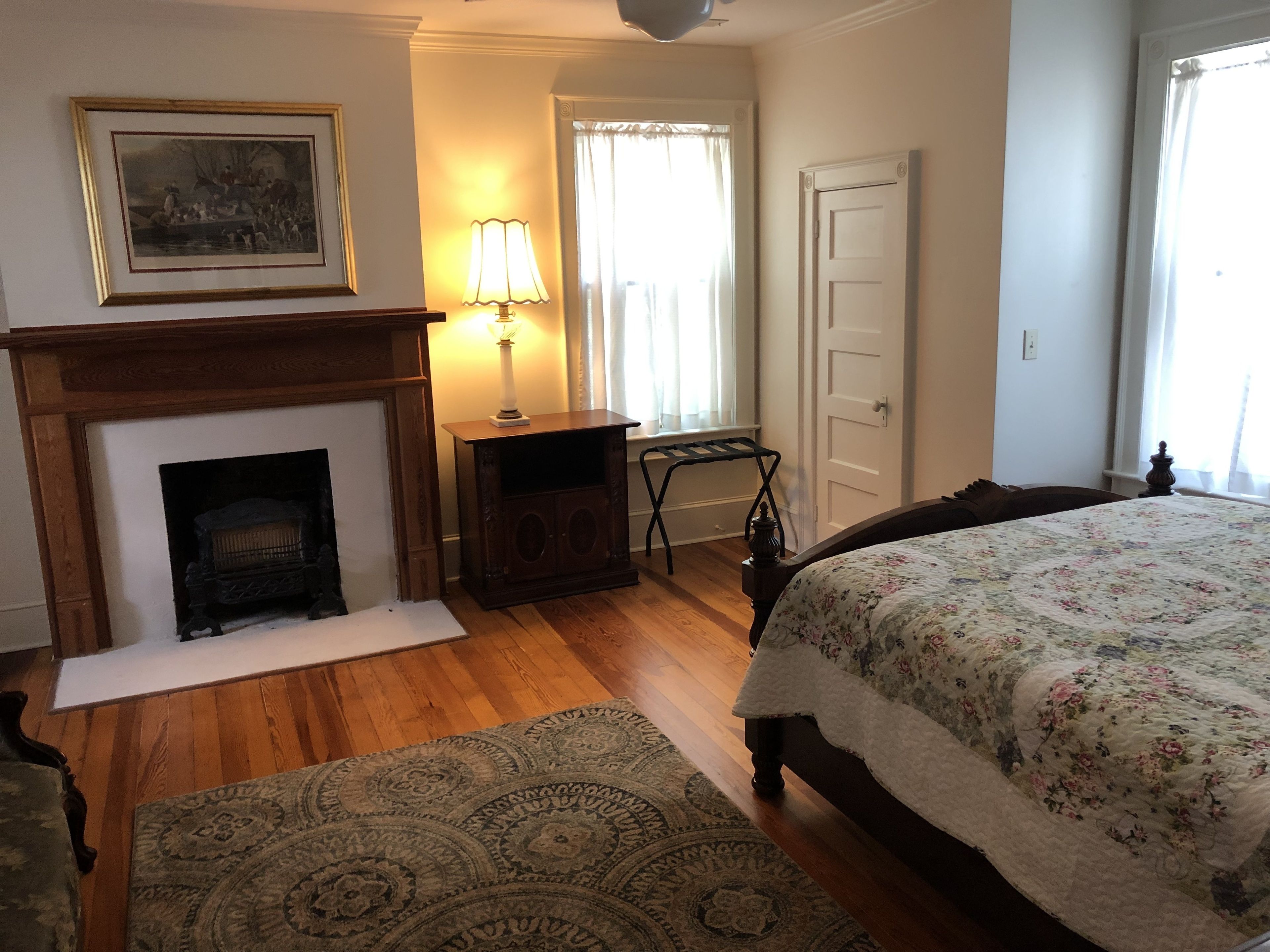 A bedroom with a fireplace, a bed with a floral bedspread, a rug, and a framed picture on the wall.