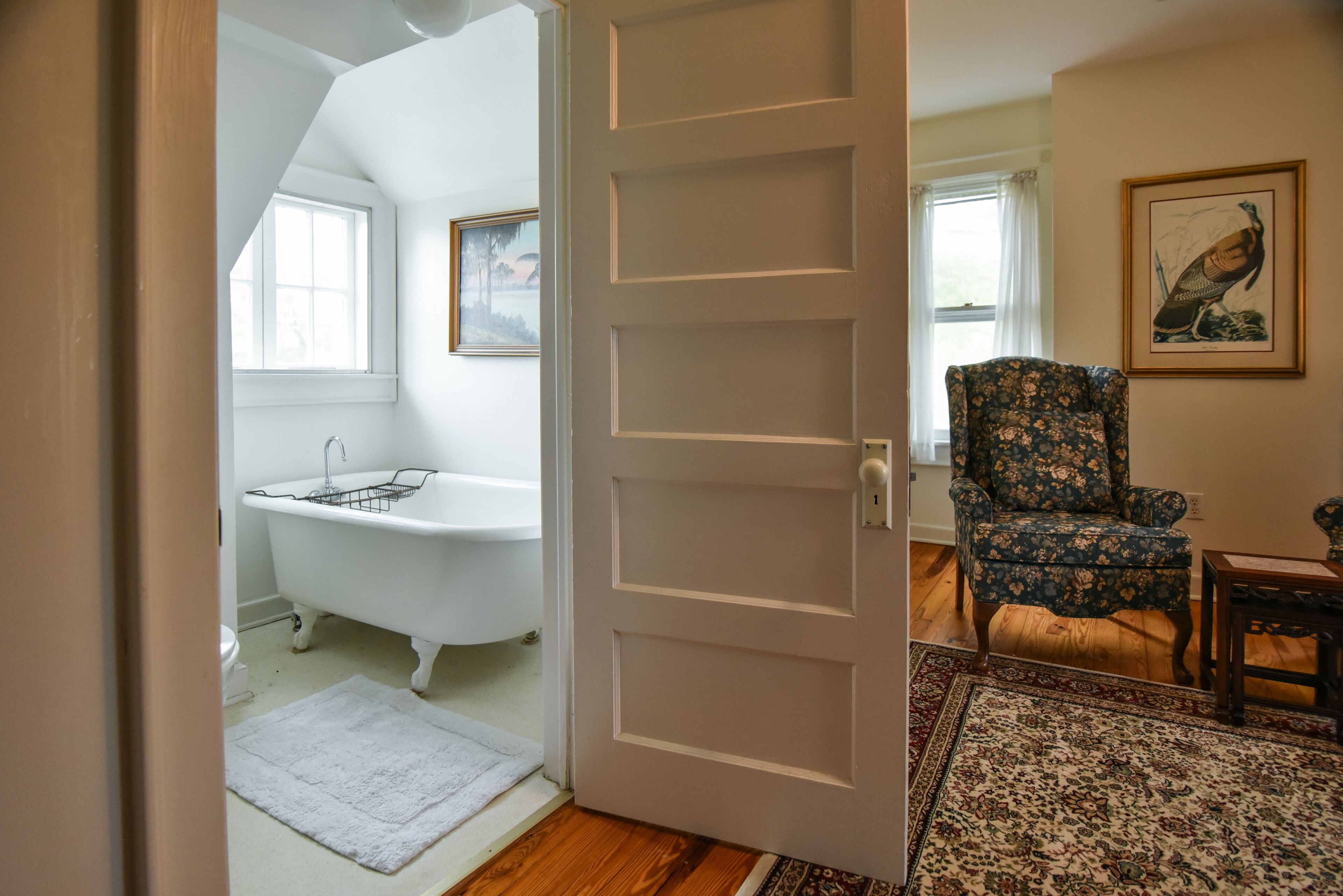 A room with a white door, a bathtub, a floral armchair, and a rug.