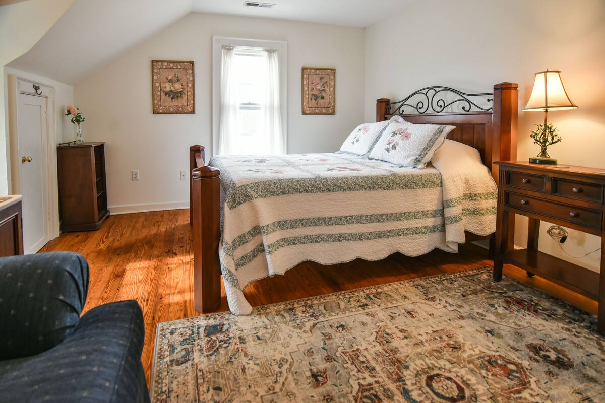 A bedroom with a wooden bed, a nightstand, a lamp, a chair, a window with curtains, and a door.