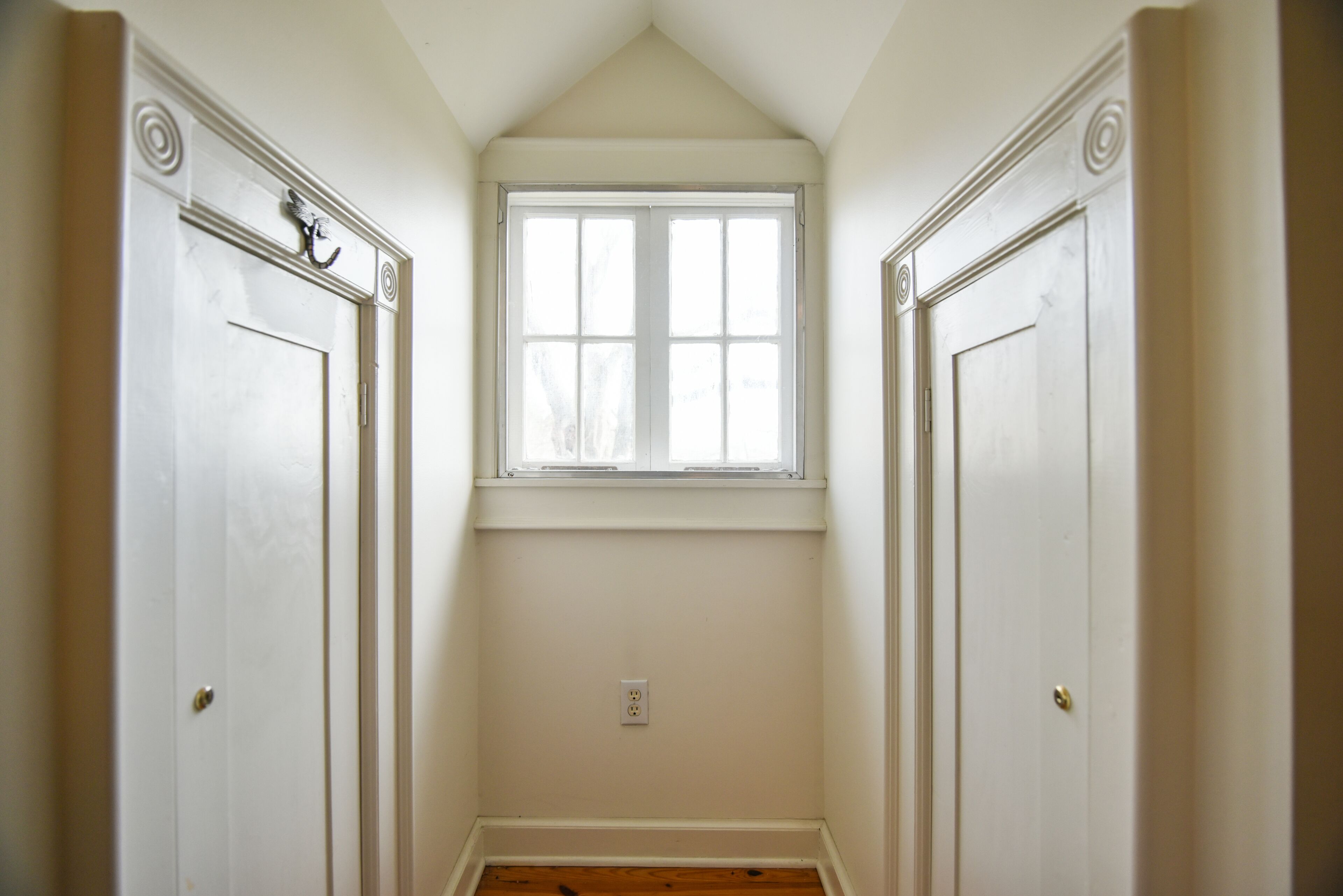 A narrow hallway with two white doors on either side, a small window at the end, and a light switch on the wall.