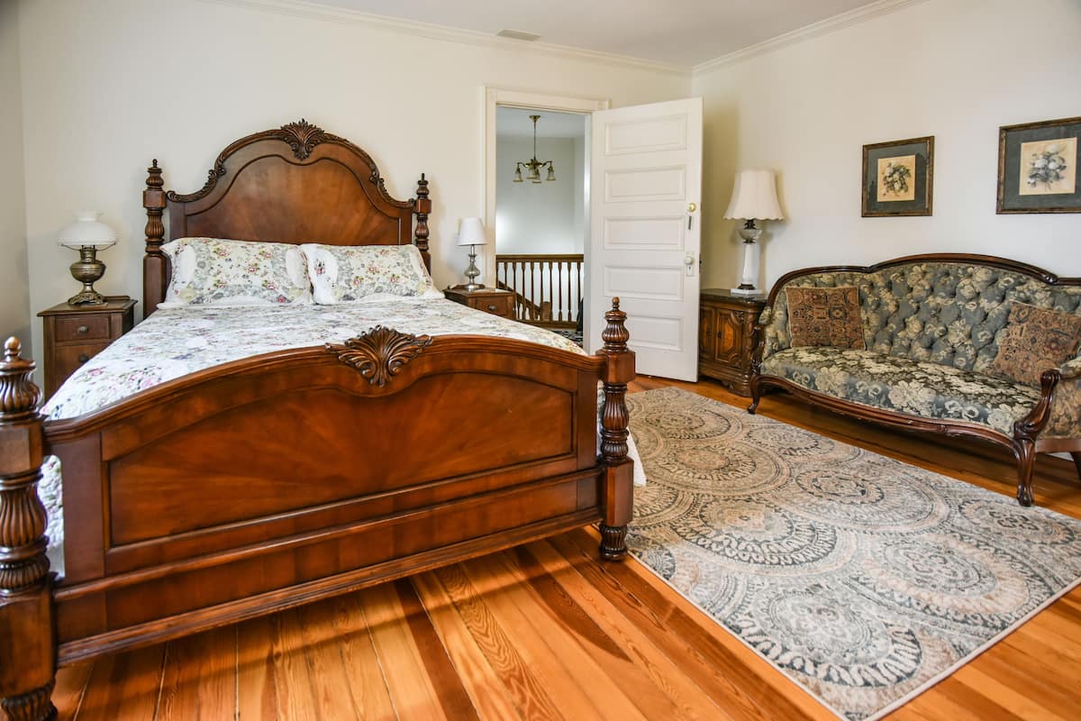 A bedroom with a wooden four-poster bed, a matching nightstand, a patterned rug, a sofa, and a door leading to another room.