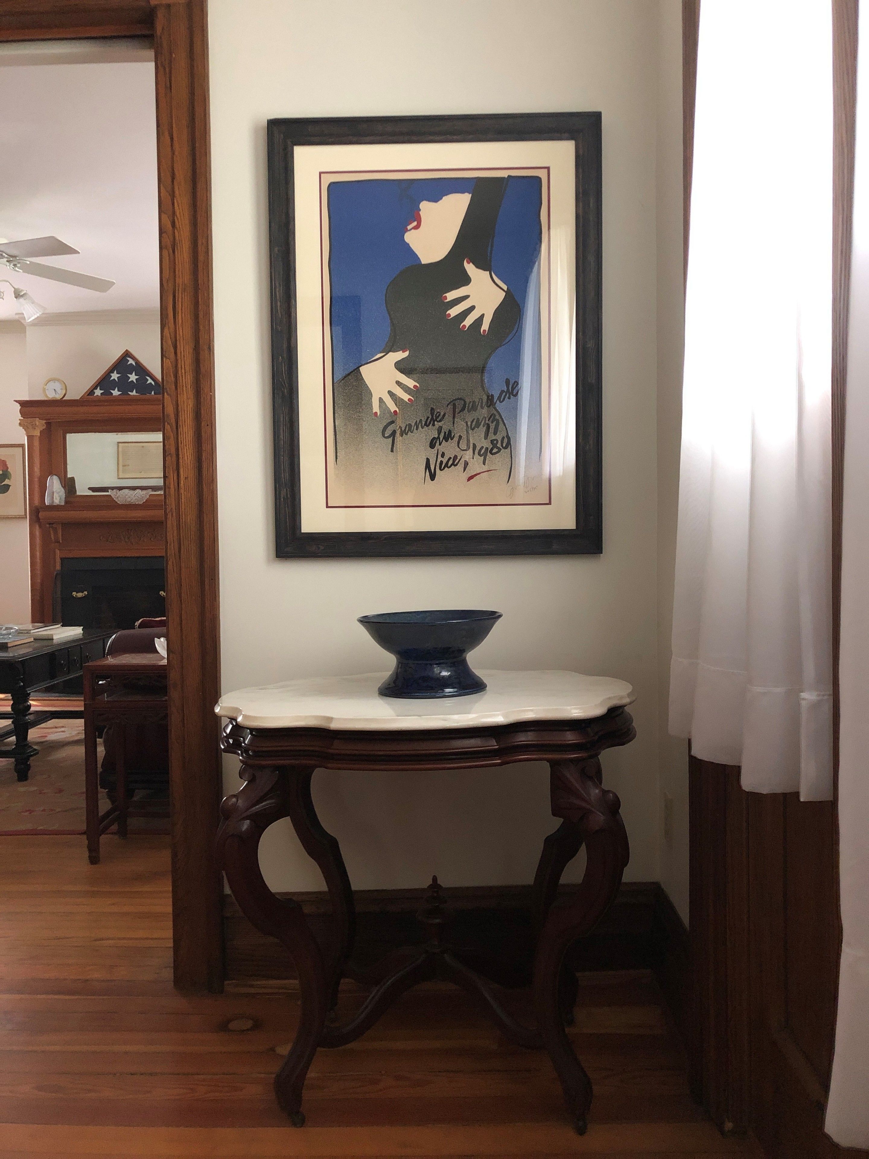 Une œuvre d’art encadrée accrochée au mur, une table avec un bol, et un plancher en bois.