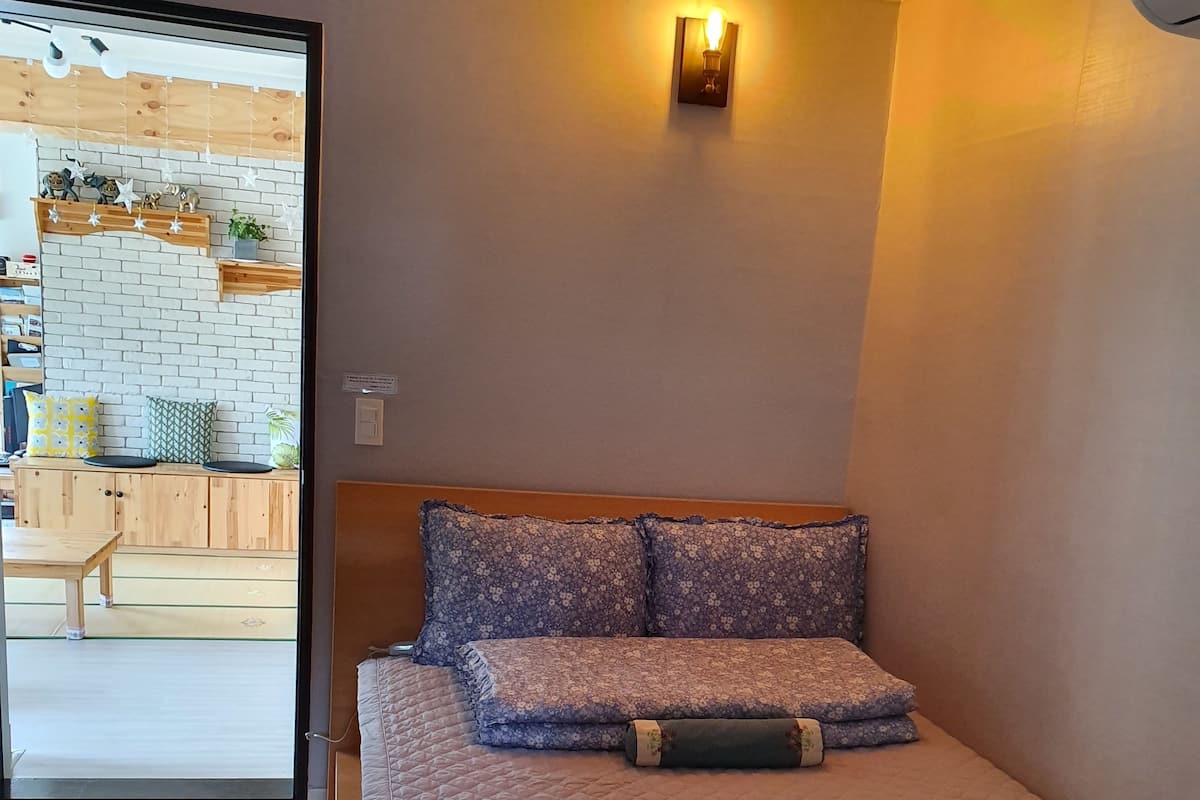 Un dormitorio con cama, mesita de noche, luz empotrada en la pared y vista a una zona de cocina.