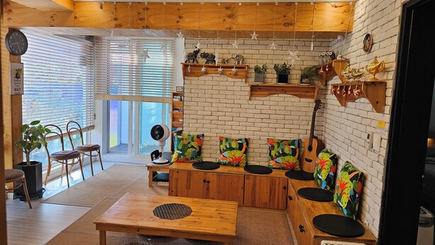 Una sala de estar acogedora con techo de madera, pared de ladrillo y un ventanal grande. Se ven dos sillones, una mesa y una guitarra.