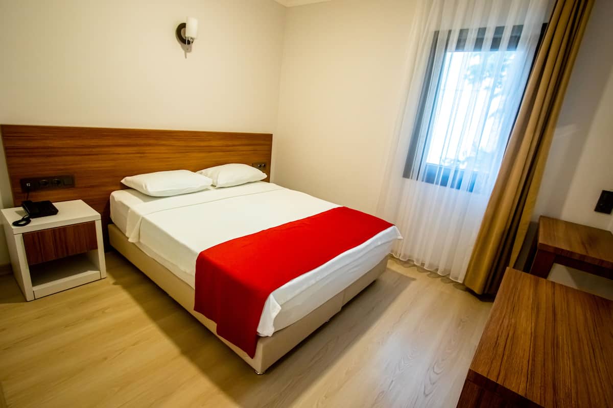 Habitació d'hotel amb un cap çal de llit de fusta, un llit blanc amb llençol vermell, una taula de nit amb telèfon i una finestra amb cortines.