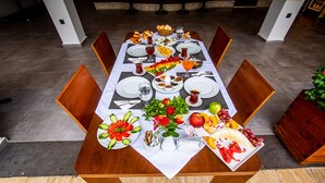 Una mesa de comedor dispuesta con diversos platos, entre ellos frutas, verduras y pan, en un interior moderno con sillas de madera y suelo de baldosas.