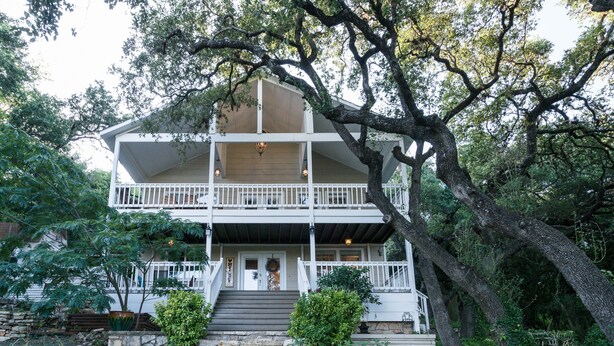 A two-story house with a balcony, surrounded by trees and a stone wall.
