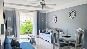 A modern living room with a sofa, dining table, and chairs. There is a ceiling fan, a window with curtains, and a view of greenery outside. - Luxe Beach Front Retreat (Ocho Rios)