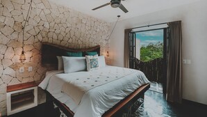 A bedroom with a stone wall, a bed with a wooden headboard, a ceiling fan, and a view of greenery through a door. - Maka Hotel Boutique (Tulum)