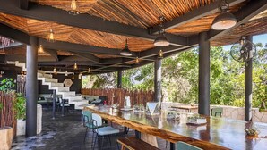 An outdoor dining area with a long wooden table, multiple chairs, and hanging pendant lights. - Maka Hotel Boutique (Tulum)