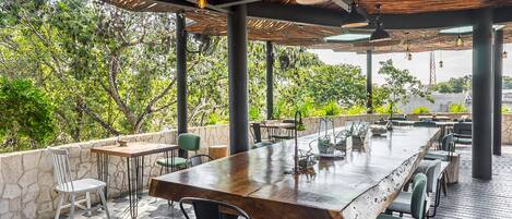 Un espacio de comedor al aire libre con una larga mesa de madera y sillas, rodeado de muros de piedra y vegetación.