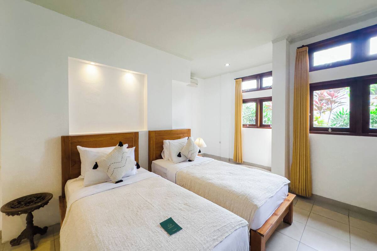 A hotel room with two single beds, a small table, a bench, and a window with curtains.