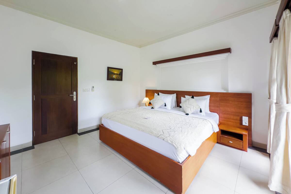 A bedroom with a wooden bed, white bedding, a nightstand, a wall-mounted shelf, and a door.