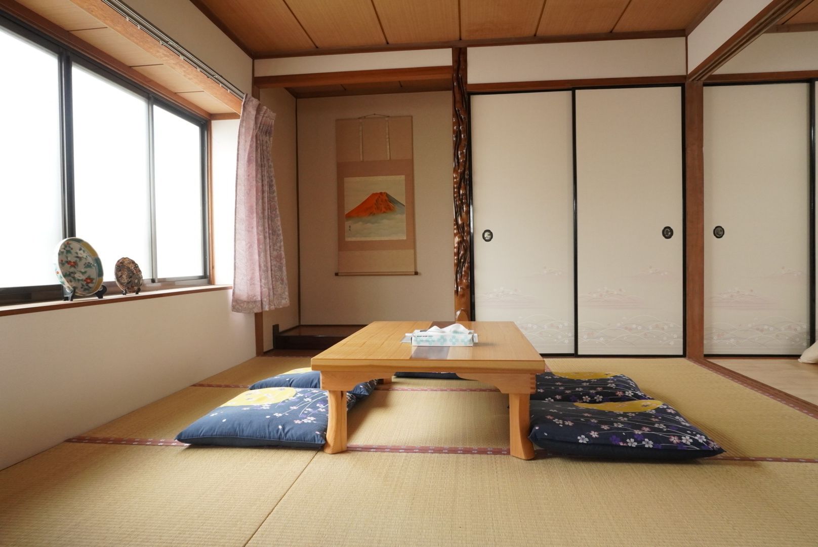 Una habitación con suelo de esteras de tatami, una mesa baja, cojines, una puerta corredera y una ventana con platos decorativos.
